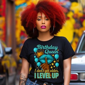 May include: Black t-shirt with the text "Birthday Queen" and the phrase "I don't get older, I LEVEL UP" in teal. The design includes a crown, lips, and balloons. The person in the image is wearing blue jeans.
