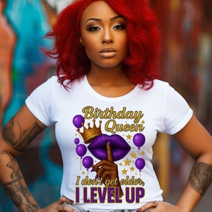 May include: White t-shirt with the text "Birthday Queen, I don't get older, I LEVEL UP" in gold and purple. The design includes a gold crown, purple lips, balloons, and stars. The person in the image has red hair and is wearing blue jeans.