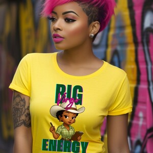 May include: A yellow t-shirt with the words "BIG Virgo ENERGY" and a cartoon cowgirl graphic. The person in the image has pink hair and is wearing black jeans. The background is a colorful graffiti wall.