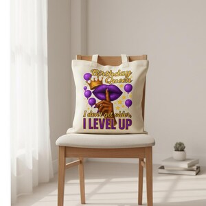 May include: A cream-colored tote bag with the text "Birthday Queen" in gold and purple, featuring a crown, lips, and balloons. The bag also has the text "I don't get older, I LEVEL UP" in purple. The bag is sitting on a wooden chair.