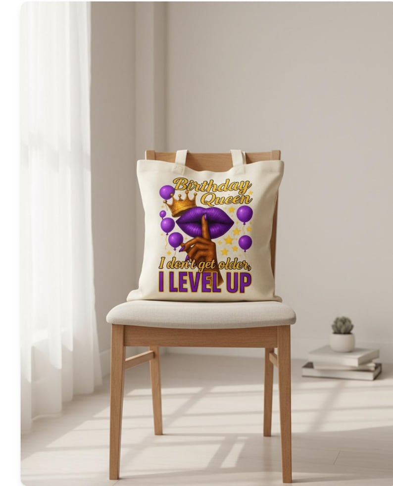 May include: A cream-colored tote bag with the text "Birthday Queen" in gold and purple, featuring a crown, lips, and balloons. The bag also has the text "I don't get older, I LEVEL UP" in purple. The bag is sitting on a wooden chair.