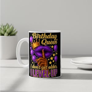 May include: White ceramic mug with a black design featuring the text "Birthday Queen" in gold and purple. The design includes a crown, purple lips, balloons, and the phrase "I don't get older, I LEVEL UP."