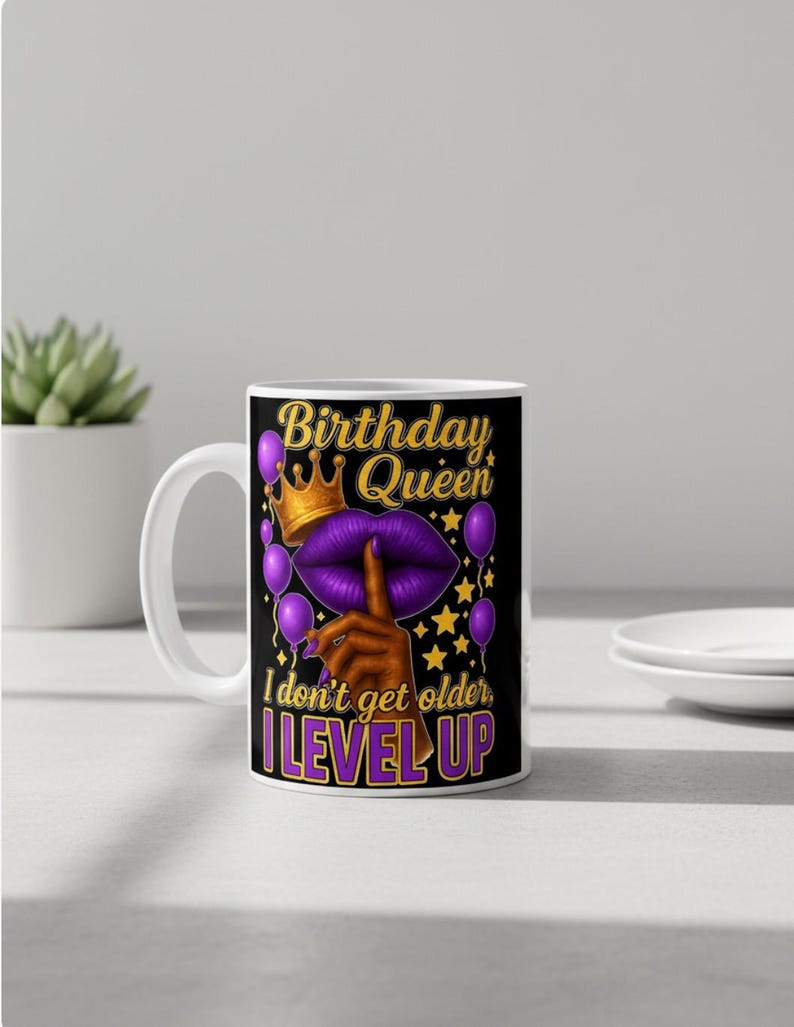 May include: White ceramic mug with a black design featuring the text "Birthday Queen" in gold and purple. The design includes a crown, purple lips, balloons, and the phrase "I don't get older, I LEVEL UP."
