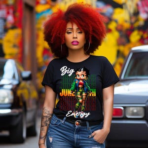May include: A woman with red hair is wearing a black t-shirt featuring a cartoon image of a woman in a green and black outfit. The t-shirt has the text "Big Juneteenth Energy" on it.