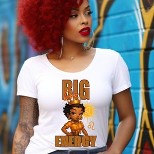 May include: White t-shirt with the text "BIG Leo ENERGY" and a cartoon illustration of a woman wearing a crown. The woman has dark skin and is wearing a gold-colored top. The background is a colorful graffiti wall.