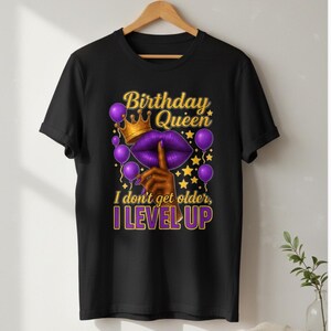 May include: Black t-shirt with a graphic design featuring the text "Birthday Queen" above a purple lip design with a gold crown and balloons. The text "I don't get older, I level up" is below the lip design.