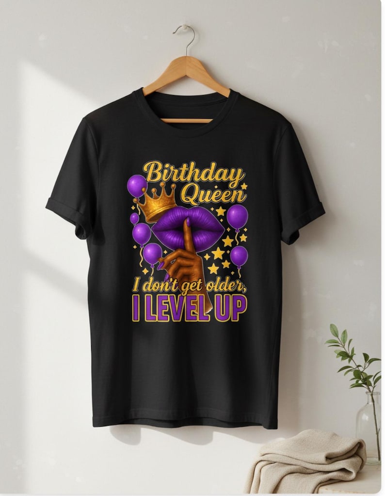May include: Black t-shirt with a graphic design featuring the text "Birthday Queen" above a purple lip design with a gold crown and balloons. The text "I don't get older, I level up" is below the lip design.