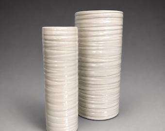 May include: Two cylindrical ceramic vases in a glossy off-white finish. Each vase features a textured surface with horizontal, raised, undulating bands that wrap around the entire body, creating a layered, organic appearance in a neutral tone.