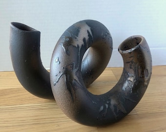 May include: A black and brown ceramic vase with a unique, twisted design. The vase has two openings, making it perfect for displaying flowers or other decorative items.