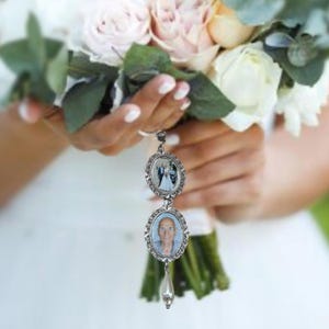 May include: A silver charm with two oval-shaped photo frames. The top frame features a photo of a couple, while the bottom frame features a photo of an older man. The charm is attached to a pearl-like bead and a silver chain.