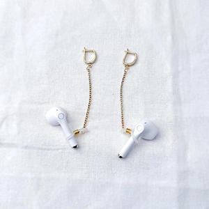 May include: A pair of white wireless earbuds with gold-coloured chain and clasp earring attachments. The earbuds have a small black tip and a circular design. The earrings are displayed on a white, textured surface.