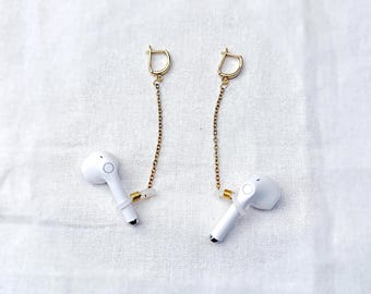 May include: A pair of white wireless earbuds with gold-colored chain and clasp earring attachments. The earbuds have a small black tip and a circular design. The earrings are displayed on a white, textured surface.