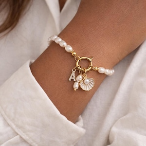 Peut inclure: Un bracelet doré orné de perles blanches et d'un charm décoratif. Le charm comprend la lettre "A" avec de petits cristaux, un pendentif coquillage et une petite perle. Le bracelet est fermé par un fermoir doré.