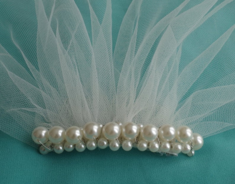 May include: A white tulle veil with a row of pearl beads attached to the bottom edge. The veil is on a turquoise background.