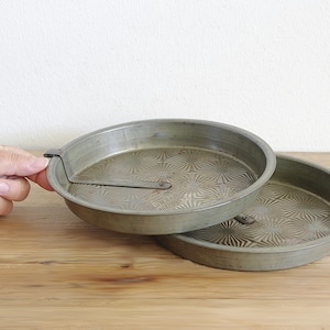 May include: Two round, metallic cake pans with a textured, starburst-like pattern on the interior base. One pan features a metal lever arm attached to the rim, designed to assist in releasing baked goods. The pans have a dull, weathered finish.