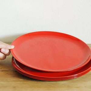 May include: A stack of red ceramic plates on a wooden surface. The top plate is being held by a hand. The plates are round and have a glossy finish. The plates are approximately 10 inches in diameter.
