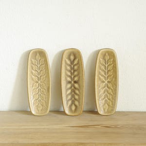 May include: Three oblong, ceramic serving dishes with a leaf design. The dishes are a light tan color and are arranged on a wooden surface. The background is a plain white wall.