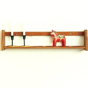 May include: Wooden shelf with two black candle holders, two white candles, and a red Dala horse figurine. The shelf is made of light brown wood and is mounted on a white wall. The horse has a decorative saddle.