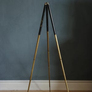 Puede incluir: Un trípode vintage con tres patas. La sección superior de cada pata es negra, pasando a un acabado dorado. El trípode se apoya sobre un suelo de madera contra una pared azul oscuro.