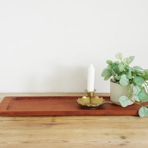 May include: A rectangular, reddish-brown wooden tray with a raised edge. A brass candle holder with a white candle and a small potted plant with green leaves sit on the tray. The tray is on a wooden surface.