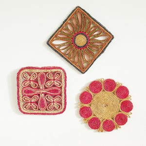May include: Three decorative jute coasters in various shapes. One is square with a red and tan butterfly design, another is diamond-shaped with a floral pattern, and the third is a round coaster with red circles around a tan centre.