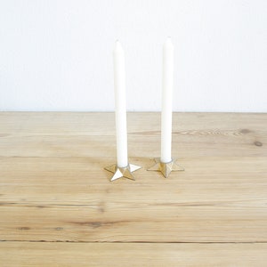 Peut inclure: Deux bougies blanches fines sont placées dans des bougeoirs dorés en forme d'étoile sur une surface en bois clair. Les bougies sont longues et minces, et les bougeoirs ont une finition texturée. Le fond est un mur blanc uni.