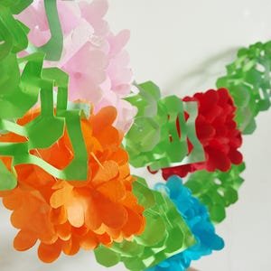 May include: A colorful paper garland featuring green, orange, pink, red, and blue floral and leaf-like cutouts. The garland is arranged in a curved shape, with the different colors creating a vibrant and festive appearance.