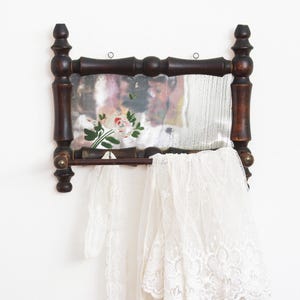 May include: An antique wall-mounted shelf with a mirror and a small shelf. The dark wood frame has decorative finials. The mirror has a floral design. A white lace curtain hangs from the shelf.