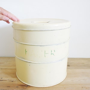 Può includere: Un contenitore vintage in metallo color crema con coperchio e fessura. Il contenitore ha tre sezioni impilate con testo verde sbiadito. Il contenitore è su una superficie di legno.