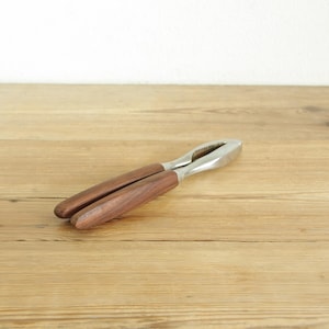 May include: A nutcracker with a stainless steel cracking mechanism and two wooden handles. The nutcracker is lying on a light brown wooden surface. The handles are a dark brown colour.