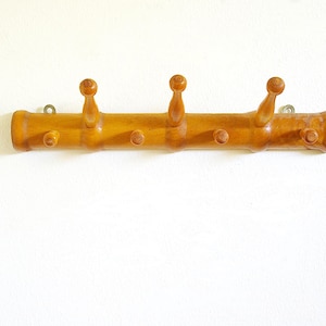 May include: A vintage wooden coat rack with a light brown finish. It features three rounded pegs and three smaller pegs, all evenly spaced along the horizontal bar. The rack is mounted on a white wall.