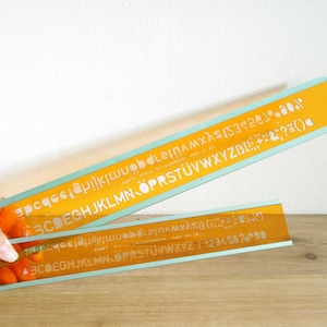 Op de afbeelding: Twee gele plastic linialen met een blauwe streep. De linialen hebben het alfabet en cijfers erop gedrukt. De linialen liggen op elkaar.