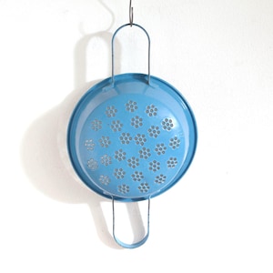 May include: A bright blue metal colander featuring a circular bowl with a series of small, flower-shaped drainage holes throughout the base. The colander includes two thin, curved metal handles attached to opposite sides of the rim for hanging.