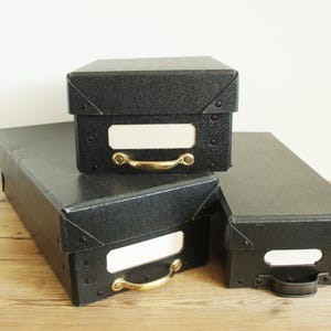 May include: Three black storage boxes with brass handles and white label holders are stacked on a wooden surface. The boxes have a textured finish and metal corner reinforcements. The boxes are rectangular and vary in size.
