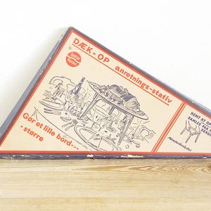 May include: A blue and white box with the text "DÆK-OP anretnings-stativ" and an illustration of a table setting. The box is for a tabletop serving stand.