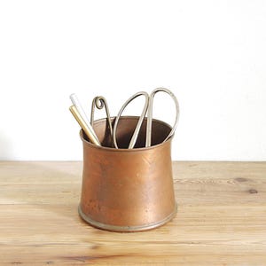 May include: A cylindrical copper container with a slightly flared base and rim. Inside the container are three metal tools: one gold-toned rod, one silver-toned rod, and a pair of silver-toned scissors with looped handles extending from the top.