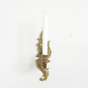 May include: A gold-colored wall sconce with a white candle. The sconce has an ornate, floral design. The candle is a standard size. The sconce is mounted on a white wall.