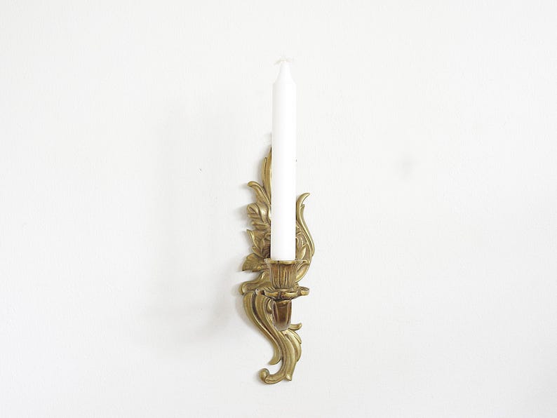 May include: A gold-colored wall sconce with a white candle. The sconce has an ornate, floral design. The candle is a standard size. The sconce is mounted on a white wall.