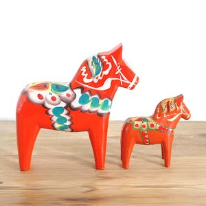 Pode incluir: Duas figuras de cavalos de madeira laranja com padrões florais e geométricos pintados à mão em branco, verde e azul. O cavalo maior está à esquerda do menor, ambos exibindo designs de sela detalhados e marcações brancas nas rédeas.