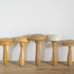 May include: Five vintage wooden hat forms in shades of light brown and tan. Each has a rounded top and a long handle. They are arranged in a row on a wooden surface against a white wall. The forms are likely used for millinery.