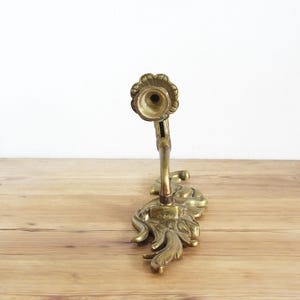 May include: An antique brass candle holder with a floral and leaf design. The candle holder has a detailed, ornate base and a curved stem leading to a circular candle cup. The brass has a warm, aged patina, and the piece is set on a wooden surface.