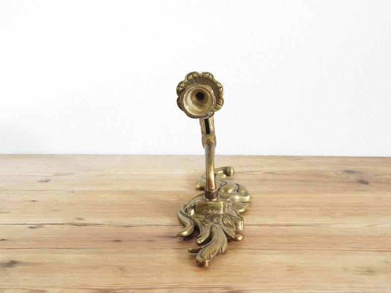 May include: An antique brass candle holder with a floral and leaf design. The candle holder has a detailed, ornate base and a curved stem leading to a circular candle cup. The brass has a warm, aged patina, and the piece is set on a wooden surface.