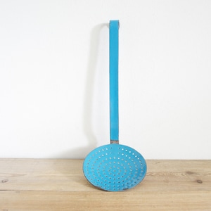 May include: A vintage turquoise metal skimmer with a long handle and a perforated round head. The kitchen utensil is designed for straining food. The skimmer is displayed against a white wall and a wooden surface.