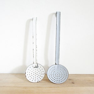 May include: Two vintage metal kitchen utensils with white and gray enamel finishes. The utensils have long handles and round perforated heads.