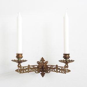 May include: An antique brass wall sconce with two white candles. The sconce features an ornate, decorative design with floral and scrollwork details. The candles are tall and slender, adding a touch of elegance to the piece.