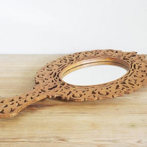 May include: A wooden hand mirror with an oval mirror and an intricately carved floral design. The handle is attached to the oval mirror. The mirror is lying on a wooden surface.