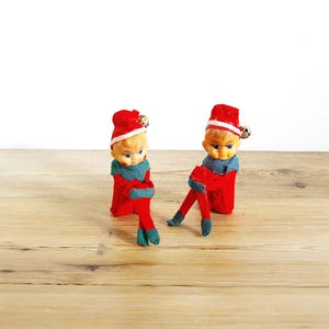 Op de afbeelding: Twee elfjes met gegoten plastic gezichten, gekleed in rode vilten mutsen met zilveren belletjes, rode vilten tunieken en donkergroene vilten kragen, manchetten en laarzen. De elfjes hebben geschilderde gelaatstrekken en zitten rechtop.