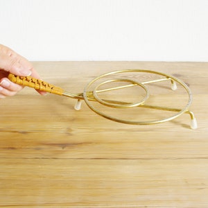 Könnte beinhalten: Ein goldfarbener Metall-Topfuntersetzer mit einem geflochtenen braunen Griff. Der Topfuntersetzer hat ein kreisförmiges Design mit drei Beinen.