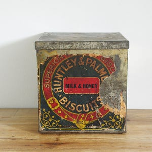 Könnte beinhalten: Vintage Huntley & Palmers Keksdose. Die rechteckige Dose hat ein abgenutztes Design mit schwarzem Hintergrund und roter und goldener Schrift. Der Text "Milk & Honey Biscuits" ist sichtbar.