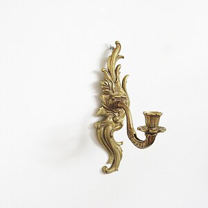 May include: Ornate brass wall sconce with a floral design. The sconce features a curved arm holding a candle cup. The decorative piece is mounted on a white wall, showcasing its intricate details and vintage aesthetic.